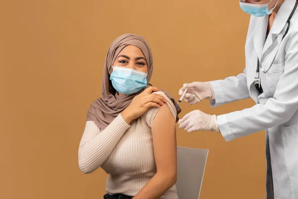 Лікар робить ін'єкції протиковзкої вакцини мусульманській жінці в масці для обличчя та хіджабі, сидячи на коричневому фоні — стокове фото