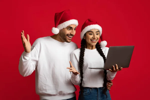 Glada unga Mellanöstern par bär Santa hattar firar framgång med laptop — Stockfoto
