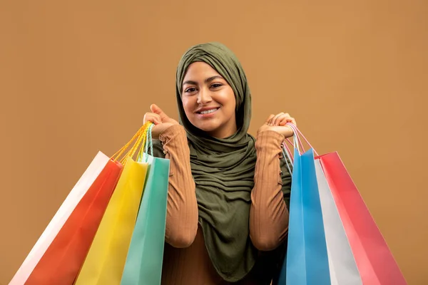 Mevsimlik satışlar. Bir sürü alışveriş çantası taşıyan tesettürlü mutlu Arap kadın kahverengi stüdyo arkaplanının başında duruyor. — Stok fotoğraf
