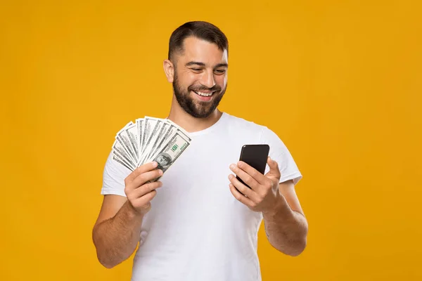 Sonriente hombre europeo de mediana edad feliz tiene muchos dólares en las manos y el teléfono inteligente —  Fotos de Stock