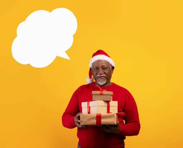 Uomo anziano di colore che indossa il cappello di Babbo Natale, regge regali di Natale, posa con bolla discorso vuoto su sfondo arancione, mockup — Foto Stock