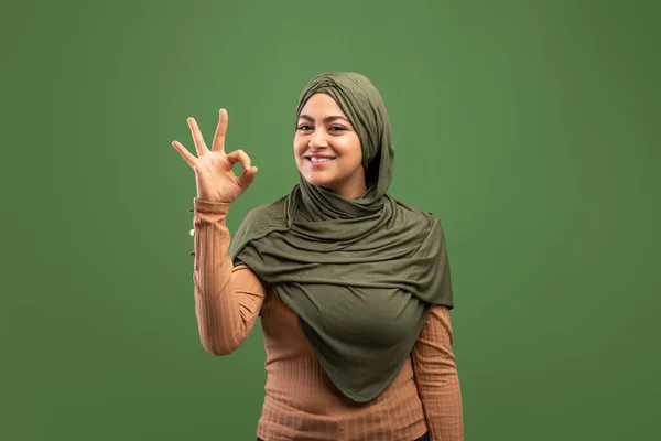 Semuanya baik-baik saja. Wanita muslim muda yang bahagia dengan kerudung menunjukkan tanda okey, berpose di atas latar belakang hijau tua — Stok Foto