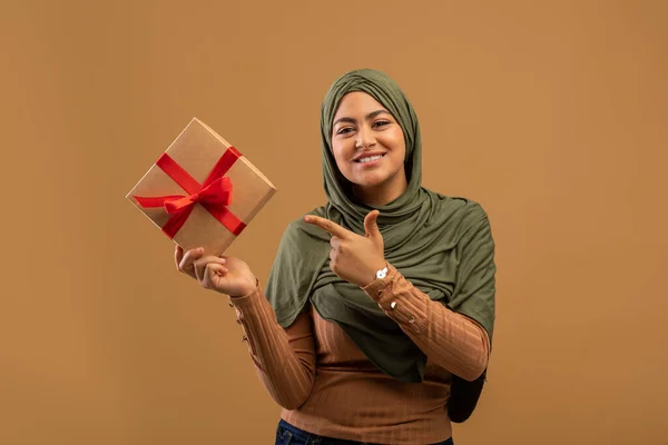 Happy arab woman holding wrapped present box and pointing finger on it, standing over brown background — Stock Photo, Image