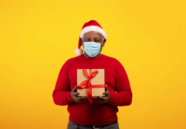 Navidad y Año Nuevo durante el bloqueo del corinavirus. Hombre negro senior con máscara protectora y caja de regalo con sombrero de Santa — Foto de Stock