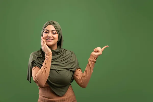 看看这个兴奋的穆斯林妇女，头戴头巾，指着自由空间，在深绿色的背景上摆出姿势，复制空间 — 图库照片