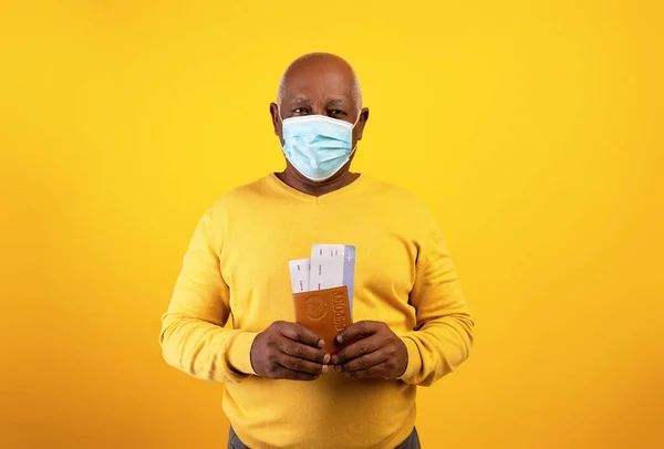 Senior svart man i medicinsk mask innehav av flygbiljetter och internationellt pass på orange studio bakgrund — Stockfoto