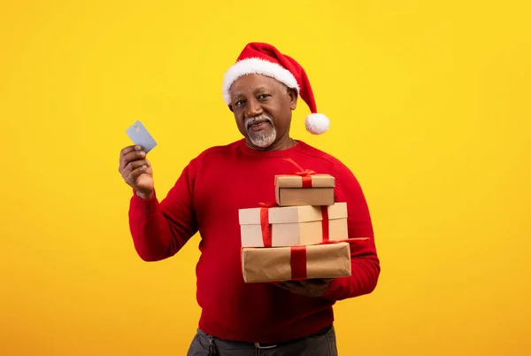 Alegre hombre negro senior en Santa Claus sombrero celebración de tarjeta de crédito y regalos de Navidad en fondo de estudio naranja — Foto de Stock