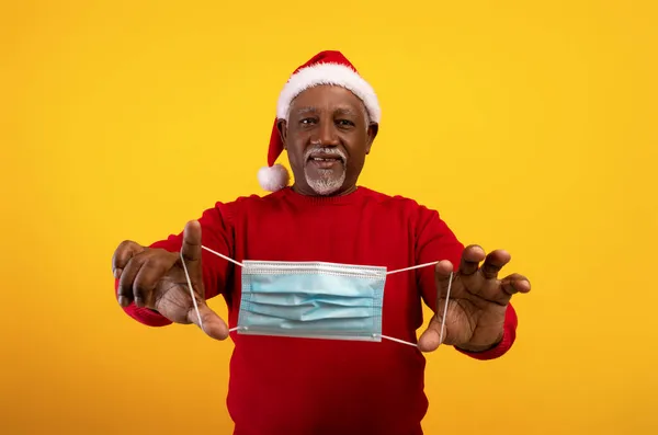 Feliz anciano negro en Santa sombrero sosteniendo la máscara en las manos extendidas, celebrando la Navidad sin coronavirus — Foto de Stock