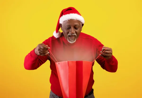 Hombre afroamericano mayor en Santa sombrero mirando dentro de la bolsa de compras con luz mágica, disfrutando de su regalo de Navidad — Foto de Stock