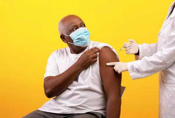 Médico haciendo vacuna contra el coronavirus a un paciente anciano negro sobre fondo de estudio naranja —  Fotos de Stock