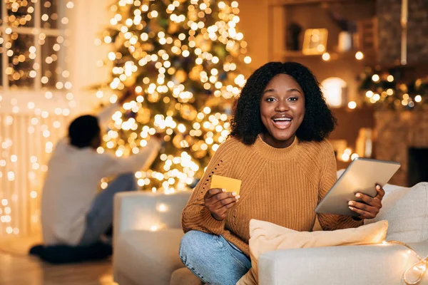 Mujer negra usando tableta y tarjeta de crédito, comprando regalos de Navidad en línea mientras su novio decora abeto en casa — Foto de Stock