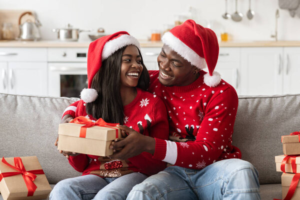 Loving millennial black couple in Santa hats exchanging xmas gifts