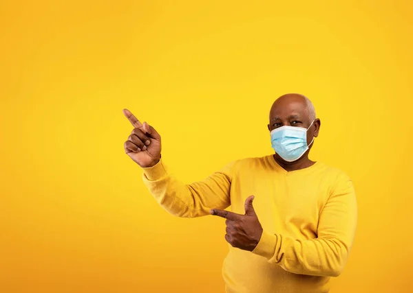 Senior svart man i skyddande ansiktsmask pekar på kopia utrymme för text eller reklam över orange bakgrund — Stockfoto