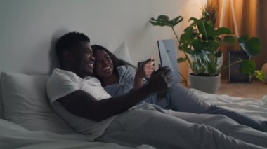Mutlu Afro-Amerikan çifti yatakta sarılıyorlar ve dijital tablette televizyon izliyorlar.