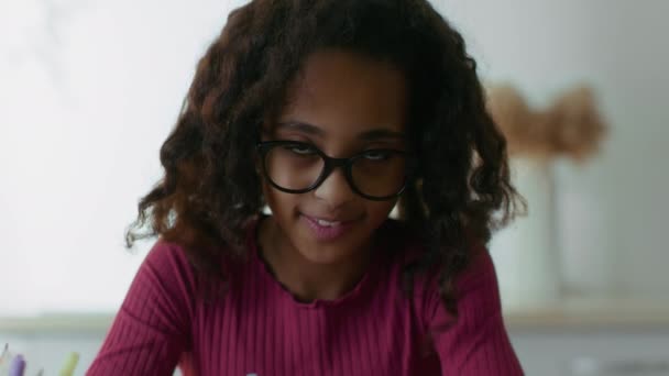 スマート小さなアフリカ系アメリカ人の女の子を身に着けています眼鏡をかけて家で宿題,頭を上げ、カメラに笑顔 — ストック動画