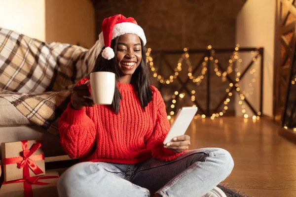 Gioiosa signora nera seduta vicino ai regali di Natale con caffè e smartphone, fare videochiamate, utilizzando l'app per lo shopping mobile — Foto Stock