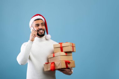 Tatille selamlama. Noel Baba şapkalı neşeli Arap adam akıllı telefondan konuşuyor ve elinde bir yığın hediye kutusu var.