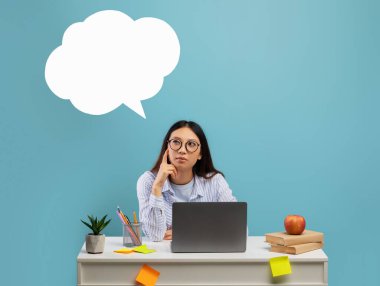 Gözlüklü düşünceli Asyalı kadın boş beyaz konuşma balonu bulutuyla düşünüyor, masada oturuyor, mavi arka planda.