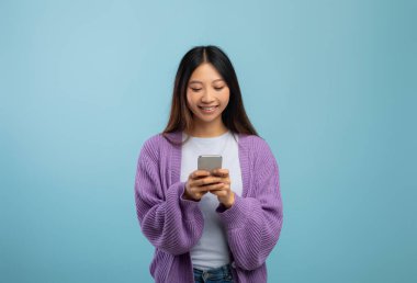 Güzel alet ve başvuru formu. Akıllı telefon kullanan, gülümseyen, mavi stüdyo geçmişi olan Asyalı bir kadın.