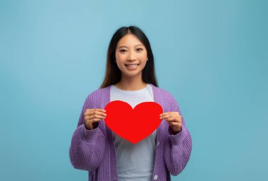 Aşk ve Sevgililer Günü konsepti. Elinde kağıt kalple gülümseyen Asyalı kadın, kameraya bakıyor ve poz veriyor, mavi arka plan