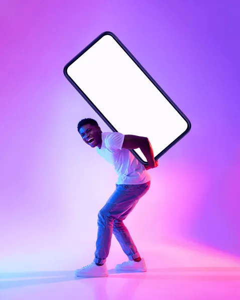 Joven negro llevando un gran smartphone pesado con pantalla vacía en la espalda con luz de neón, de cuerpo entero. Burla. —  Fotos de Stock