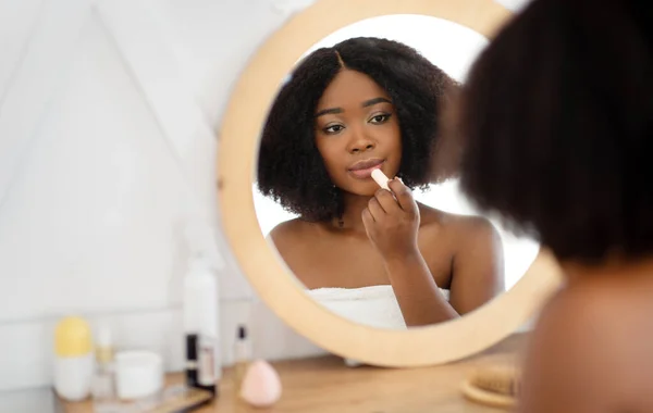 Νεαρή μαύρη κυρία να βάζει κραγιόν μπροστά από τον καθρέφτη στο σπίτι, να βάζει διακοσμητικό μακιγιάζ. Καθημερινές ρουτίνες ομορφιάς — Φωτογραφία Αρχείου