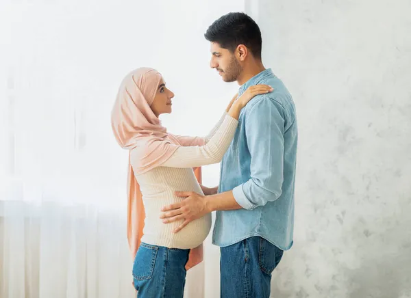 Feliz pareja musulmana esperando al bebé abrazándose y parándose juntos contra la ventana en casa, vista lateral — Foto de Stock