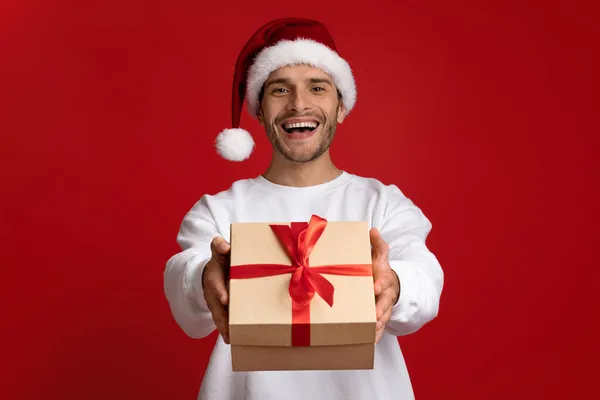 Geschenk für Sie. Aufgeregter junger Mann mit Nikolausmütze überreicht Weihnachtsgeschenk vor laufender Kamera — Stockfoto