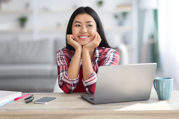 Sonhador sorrindo mulher asiática sentada na mesa com laptop e olhando para fora — Fotografia de Stock