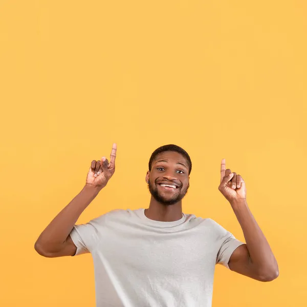 Overblijde zwarte man wijzend vingers omhoog naar de vrije ruimte en glimlachend naar de camera, staande over gele achtergrond — Stockfoto