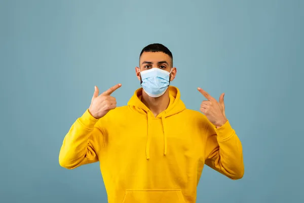 Conceito de proteção. Jovem árabe cara apontando os dedos para máscara protetora, de pé sobre fundo estúdio azul — Fotografia de Stock