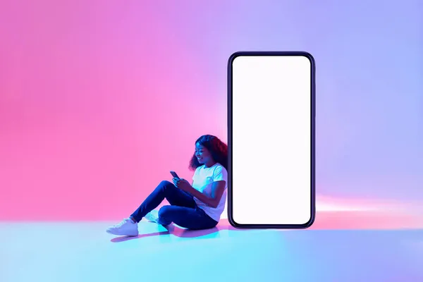 Heureuse femme noire assise près d'un grand téléphone portable avec maquette pour votre application à l'écran, en utilisant un appareil mobile dans la lumière au néon — Photo