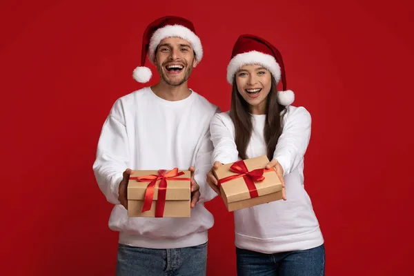 サンタの帽子をかぶっている幸せな若いカップルは、カメラでクリスマスギフトを示す — ストック写真