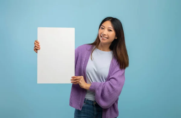 Веселая азиатка держит пустую доску для рекламы или текста, стоя на синем фоне студии — стоковое фото