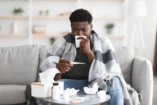 Nemocný černoch sedí na gauči, měří tělesnou teplotu — Stock fotografie