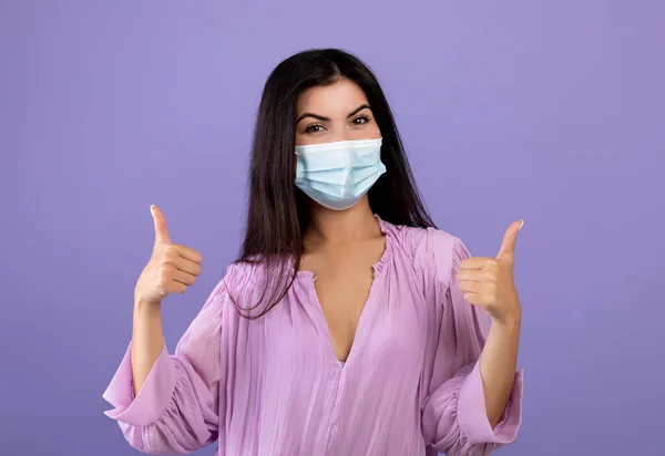Портрет армянской женщины в медицинской маске и жесты большие пальцы вверх, позируя перед камерой на фиолетовом фоне — стоковое фото