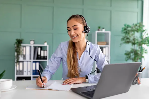 Moderne Technologie und E-Learning. Glückliche Frau mit Kopfhörer macht sich Notizen, während sie das Webinar am Laptop verfolgt — Stockfoto