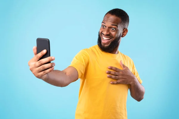 Емоційний молодий чорний хлопець бере селфі на смартфон, маючи онлайн відеочат на фоні синьої студії — стокове фото
