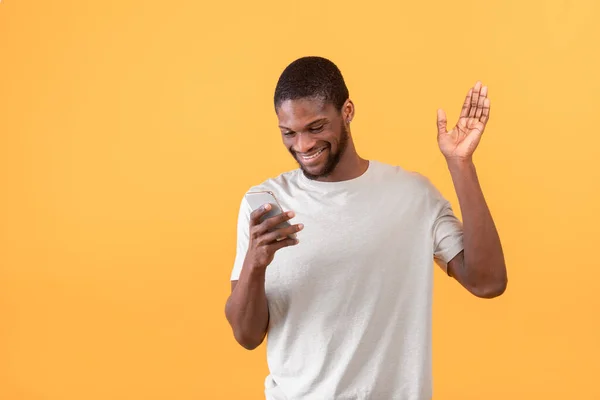 Felice uomo africano americano con smartphone fare videochiamate, agitando la mano alla fotocamera del cellulare su sfondo giallo — Foto Stock