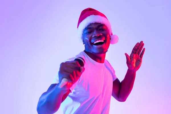 Weihnachtsmusik. Junger schwarzer Mann mit Weihnachtsmütze singt Karaoke ins Mikrofon, tanzt und amüsiert sich im Neonlicht — Stockfoto