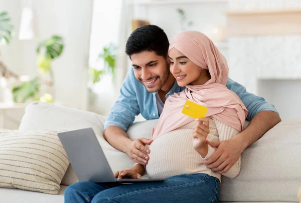 Áruk online vásárlása. Boldog muszlim terhes pár laptoppal és hitelkártyával, kanapén ülve — Stock Fotó