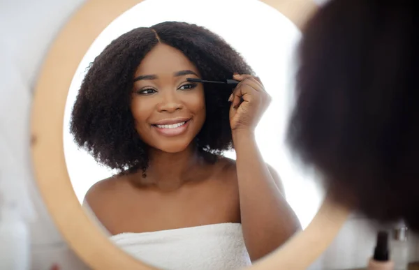 Potret wanita kulit hitam muda yang gembira mengenakan maskara setelah mandi atau mandi, melihat ke cermin di rumah. Kecantikan makeup — Stok Foto