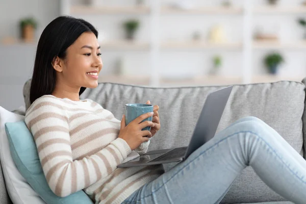 Huiselijke recreatie. jong aziatische vrouw ontspannen met koffie en laptop thuis — Stockfoto