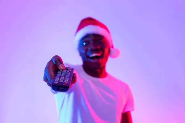 Karácsonyi mozi. Boldog fiatal fekete srác Mikulás kalap segítségével TV távirányító, néz karácsonyi filmet neon fény — Stock Fotó