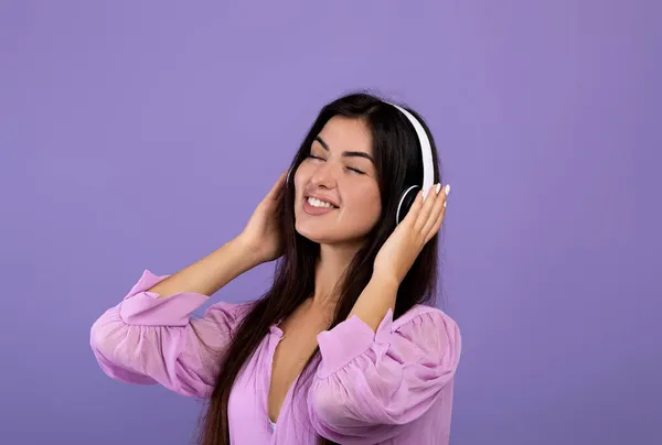 Mulher arménia despreocupada ouvindo música com olhos fechados e sorrindo, usando fones de ouvido sem fio, fundo roxo — Fotografia de Stock