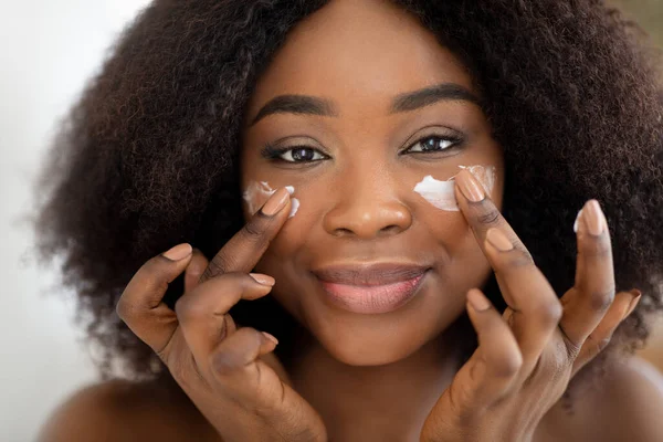 Retrato de cerca de una atractiva dama negra milenaria que aplica crema facial bajo sus ojos. Belleza y cosmética natural —  Fotos de Stock
