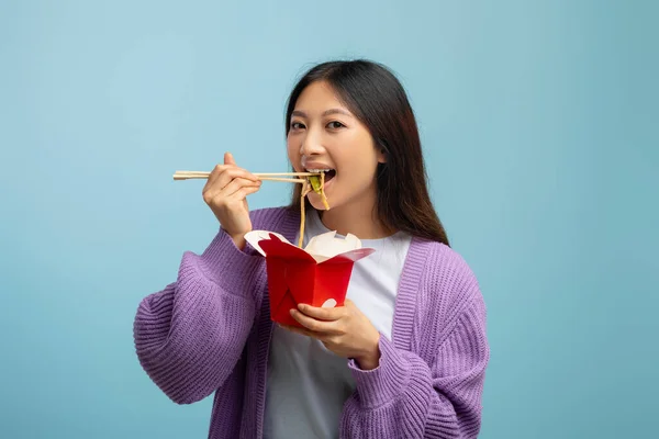 Joven mujer asiática comiendo fideos con palillos, sosteniendo la caja de papel y mirando a la cámara, fondo azul —  Fotos de Stock
