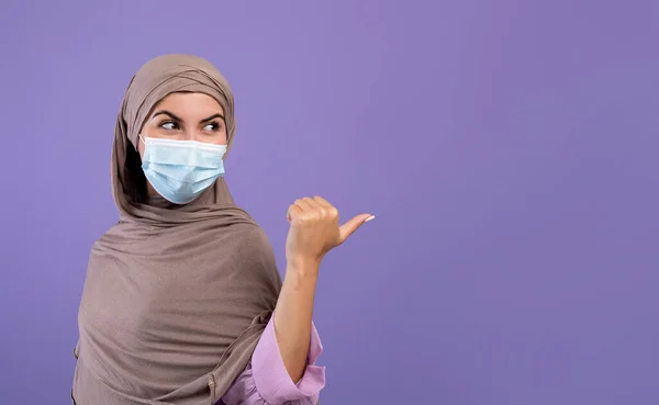 얼굴을 보호하는 마스크를 쓴 이슬람 여성 과 히잡은 보라색 배경 위에 본문이나 광고를 위한 복사 공간을 가리키고 있다 — 스톡 사진