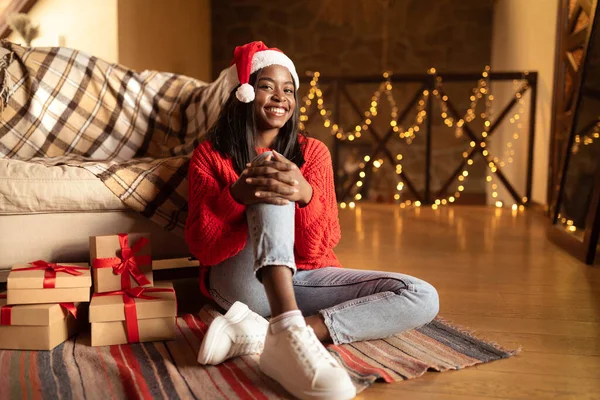 Hermosa mujer negra joven en Santa sombrero y suéter celebrando la Navidad con regalos en casa, espacio para copiar — Foto de Stock