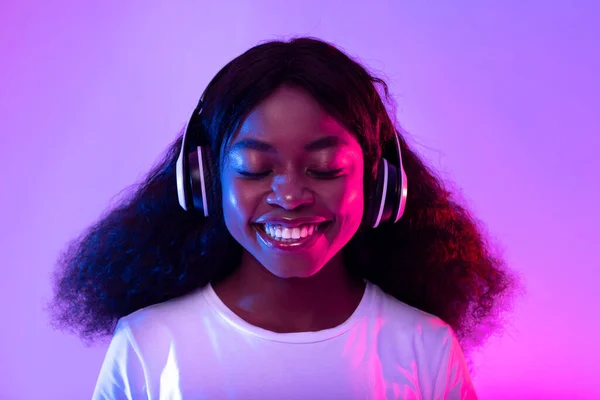Hermosa dama negra milenaria escuchando música en auriculares, sonriendo con los ojos cerrados en luz de neón — Foto de Stock